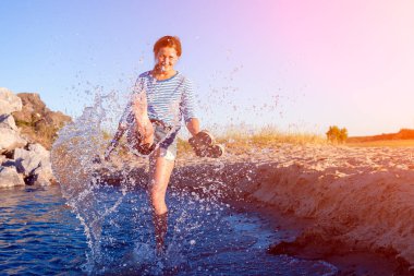 Kot pantolon ve çizgili tişört gülümsüyor, neşeli bir esmer kadın plaj boyunca yürüyor, dalgalar başladı, devamlı spreyler ve parlak güneşin bir yaz gününde sahiptir. Deniz ve canlı stil yaz tatilleri kavramı