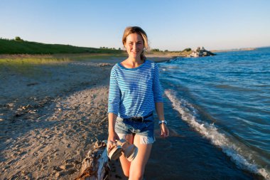 Kot pantolon ve çizgili tişört gülümsüyor, neşeli bir esmer kadın plaj boyunca yürür ve parlak güneşin bir yaz gününde sahiptir. Deniz ve canlı stil yaz tatilleri kavramı