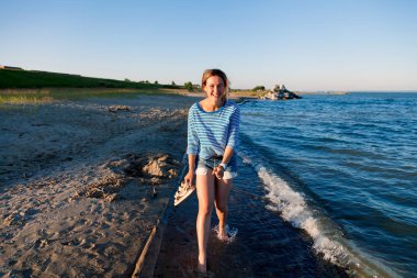 Kot pantolon ve çizgili tişört gülümsüyor, neşeli bir esmer kadın plaj boyunca yürür ve parlak güneşin bir yaz gününde sahiptir. Deniz ve canlı stil yaz tatilleri kavramı