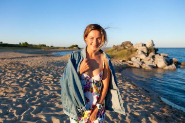 Renkli iplik ve denim ceket gülümsüyor, neşeli bir esmer kadın plaj boyunca yürür ve parlak güneşin bir yaz gününde sahiptir. Deniz ve canlı stil yaz tatilleri kavramı