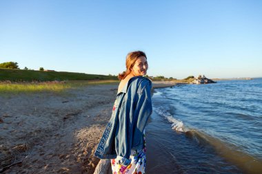 Renkli iplik ve denim ceket gülümsüyor, neşeli bir esmer kadın plaj boyunca yürür ve parlak güneşin bir yaz gününde sahiptir. Deniz ve canlı stil yaz tatilleri kavramı