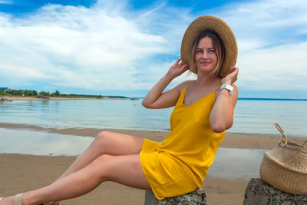 軽いカジュアルな服装と休憩とポーズを夏の日に青い空 青い空と美しい海の近くの砂浜のビーチの岩に麦わら帽子の若い美しい女性 — ストック写真