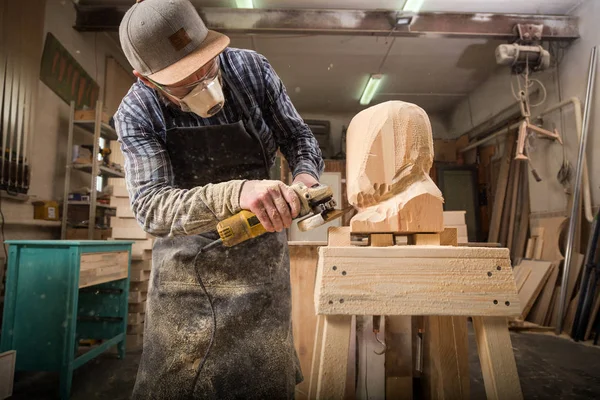 Carpenter in work clothes and small buiness owner working in woodwork workshop, processes the board with an angle grinder , on the table is a hammer and many tools