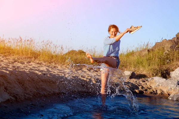 Kot pantolon ve çizgili tişört gülümsüyor, neşeli bir esmer kadın plaj boyunca yürüyor, dalgalar başladı, devamlı spreyler ve parlak güneşin bir yaz gününde sahiptir. Deniz ve canlı stil yaz tatilleri kavramı