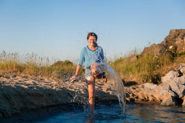 Kot pantolon ve çizgili tişört gülümsüyor, neşeli bir esmer kadın plaj boyunca yürüyor, dalgalar başladı, devamlı spreyler ve parlak güneşin bir yaz gününde sahiptir. Deniz ve canlı stil yaz tatilleri kavramı