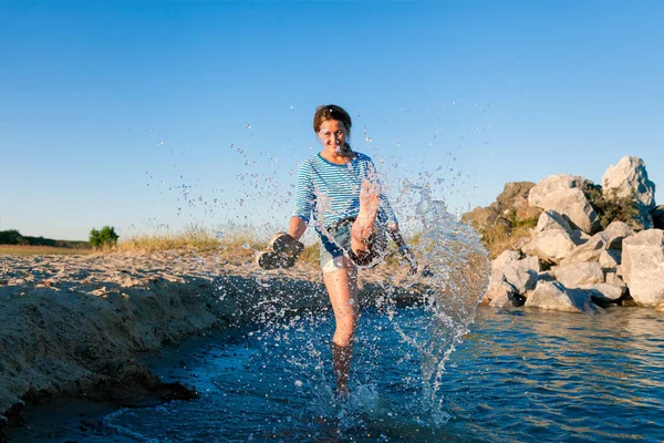 Kot pantolon ve çizgili tişört gülümsüyor, neşeli bir esmer kadın plaj boyunca yürüyor, dalgalar başladı, devamlı spreyler ve parlak güneşin bir yaz gününde sahiptir. Deniz ve canlı stil yaz tatilleri kavramı