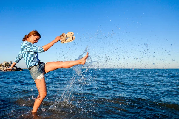 Kot pantolon ve çizgili tişört gülümsüyor, neşeli bir esmer kadın plaj boyunca yürüyor, dalgalar başladı, devamlı spreyler ve parlak güneşin bir yaz gününde sahiptir. Deniz ve canlı stil yaz tatilleri kavramı