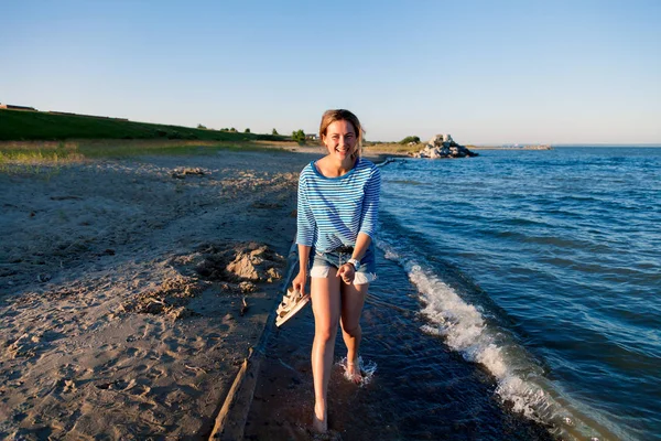 Kot pantolon ve çizgili tişört gülümsüyor, neşeli bir esmer kadın plaj boyunca yürür ve parlak güneşin bir yaz gününde sahiptir. Deniz ve canlı stil yaz tatilleri kavramı