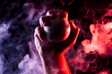 Close-up of a strong male hand holding a white baseball ball  against a background of colored  blue  and red smoke and black isolated background.  clipart