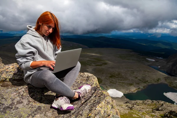 Young tourist woman working on a laptop and sitting on the top of the mounting and looking at a beautiful  landscape.  Freelancer works outside the office. The concept of traveling and working outside the office
