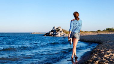 Kot pantolon ve çizgili tişört gülümsüyor, neşeli bir esmer kadın plaj boyunca yürür ve parlak güneşin bir yaz gününde sahiptir. Deniz ve canlı stil yaz tatilleri kavramı