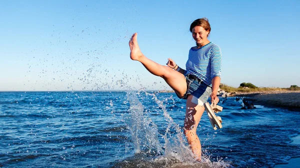 Concept of summer holidays at sea and live style. A young dark-haired woman smiles, walks along the beach, play with waves, sprays around a lot and enjoys the bright sun on a summer day.