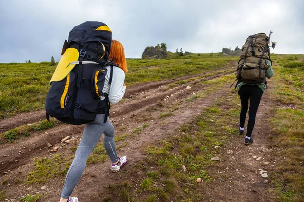 Travel Lifestyle and survival concept rear view. Hiking women  with backpack  enjoying the trekking, go to the mountains,  in the background a green field, mountains. The sun shines, the blue sky on a warm summer day.