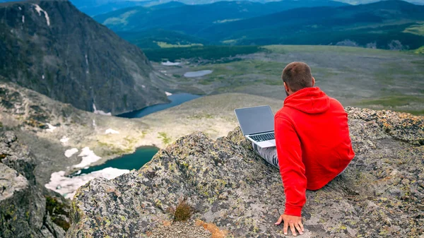 Young tourist man working on a laptop and sitting on the top of the mounting and looking at a beautiful  landscape.  Freelancer works outside the office. The concept of traveling and working outside the office