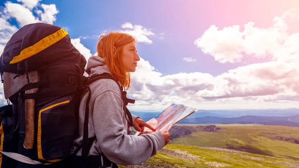 Stylish woman with backpack hiking, orient themselves to the terrain, study map and navigate the route through the mountains. Travel Lifestyle and survival concept rear view. Hipster traveler on top of mountains