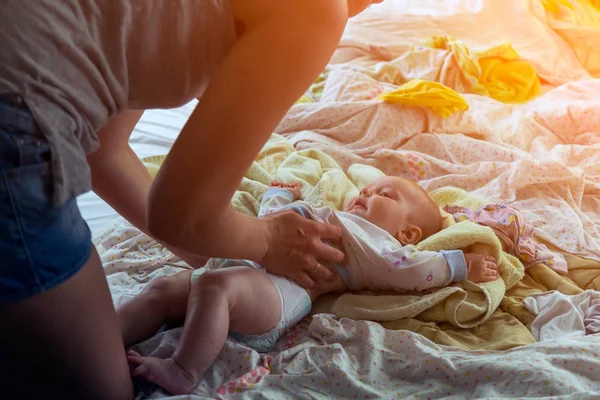 A close-up of a young slender mother swaddles and dresses a little baby girl on a light bed. Home obligations of a young mother — Stock Image Close Young Slender Mother Swaddles Dresses Little Baby Girl Light Stock Image