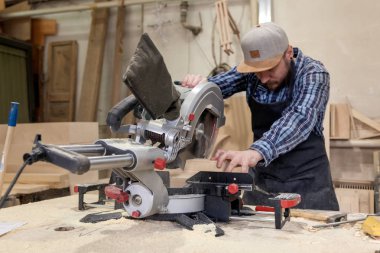 İş elbiseleri ve doğrama atölyesinde çalışan küçük buiness sahibi arasında masadaki bir ahşap tahta kesmek için bir daire testere kullanarak deneyimli marangoz bir çekiç ve birçok araç olduğunu