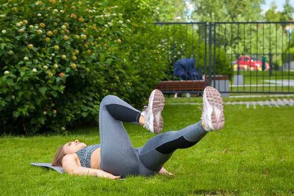 A beautiful thin woman sportswoman doing an exercise on the development of the gluteal muscles, hip bridge exercise on the rug for yoga before training on a green lawn in the park on a summer day