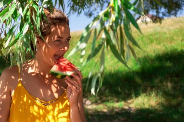 Mutlu gülen, gülümseyen ve tropikal altında bir karpuz yeme genç güzel kadın arka plan yeşil çim ve plaj içinde yeşil ağaç yaprakları