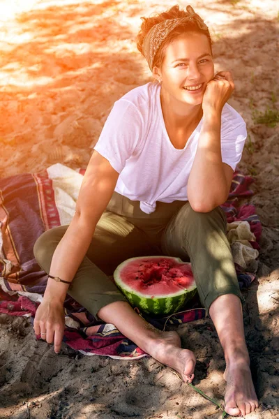 Young beautiful woman laughs cheerfully, smiles, holds between the legs a big watermelon and eats, sitting on a sandy beach in a summer day near the sea — Stock Image Young Beautiful Woman Laughs Cheerfully Smiles Holds Legs Big Watermelon Royalty Free Stock Images