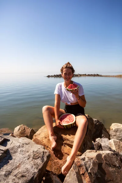 Young beautiful woman laughing happily, smiling, holding between the legs a big watermelon and eating, sitting stone rocks on a summer day near the sea — Stock Image Young Beautiful Woman Laughing Happily Smiling Holding Legs Big Watermelon Royalty Free Stock Images