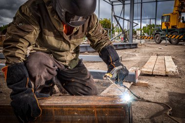İn yakın güçlü bir kaynakçı kahverengi üniforma, kaynak maskesi ve kaynakçılar deri biri, metal bir ürün ile bir ark kaynak makinesi inşaat sahasında kaynaklı, mavi kıvılcımlar taraf için