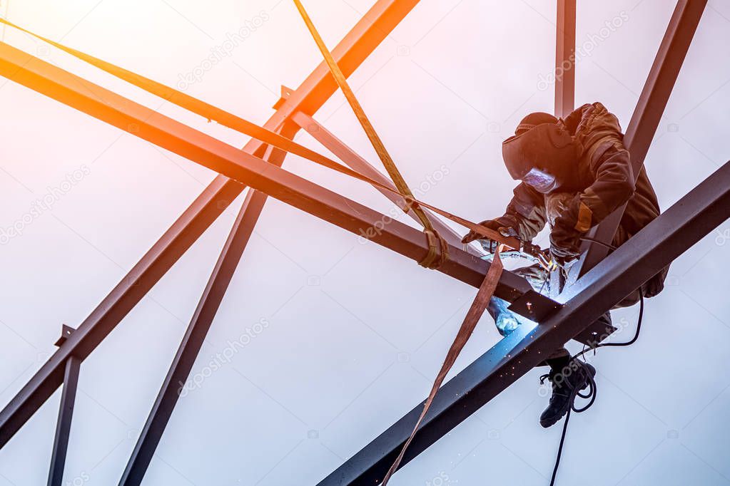 A working man is an engineer and welder in a construction overall, a welding mask is cooking metal and is sitting on a metal structure at an altitude against the blue sky
