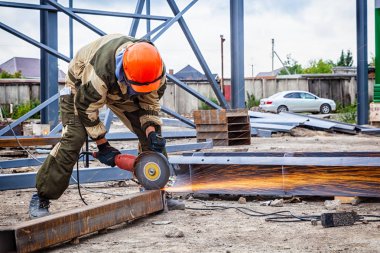 A young  man welder in brown uniform, a construction helmet and welders leathers, grinder metal an angle grinder  at the construction site, orange sparks fly to the sides clipart