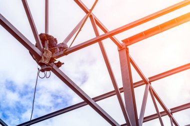 Çalışan bir adam bir mühendis ve kaynakçı bir inşaat genel olarak, bir kaynak maskesi metal yemek ve bir metal yapı mavi gökyüzüne karşı bir irtifada oturuyor