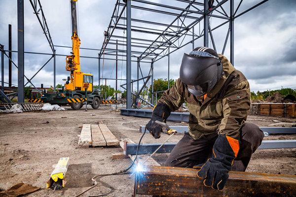 A young  man welder in brown uniform, welding mask and welders leathers, weld  metal  with a arc welding machine at the construction site, blue sparks fly to the sides