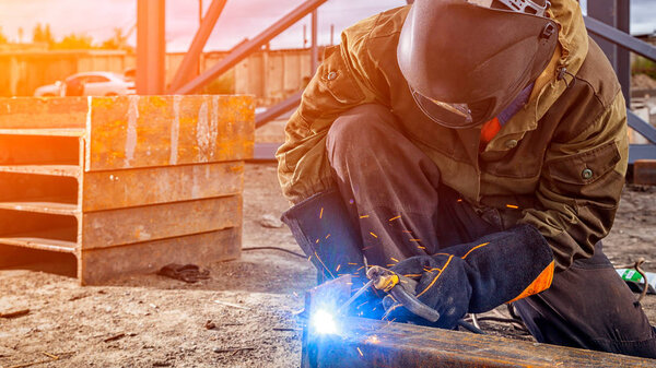 A young  man welder in brown uniform, welding mask and welders leathers, weld  metal  with a arc welding machine at the construction site, blue sparks fly to the sides