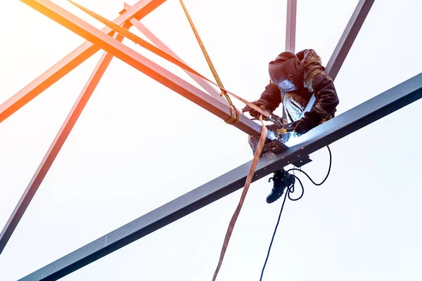 A man  welder working at height with insurance  in  welding mask is weld metal and is sitting on a metal structure at an altitude against the blue sky