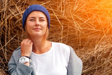Güneşin haystacks arka planı açık havada yürüyüş ve bir kat, örgü şapka, kot giyen oldukça taze darkhaired genç kadın. Sonbahar tatil köyü ve canlı stil de kavramı