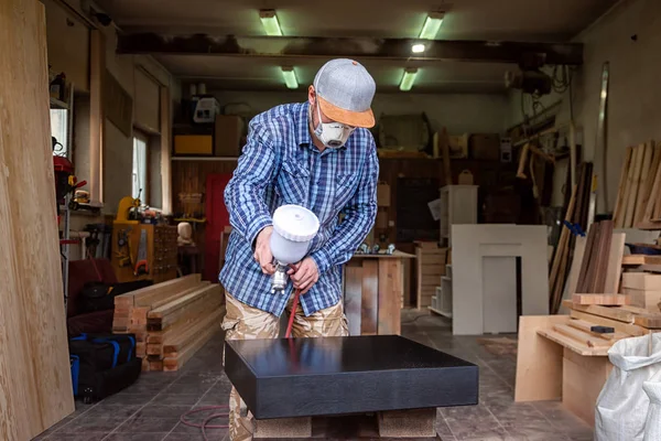 Experienced carpenter in work clothes and small buiness owner paints a wooden box from the dresser in black color in workshop, in the background a lot of tools