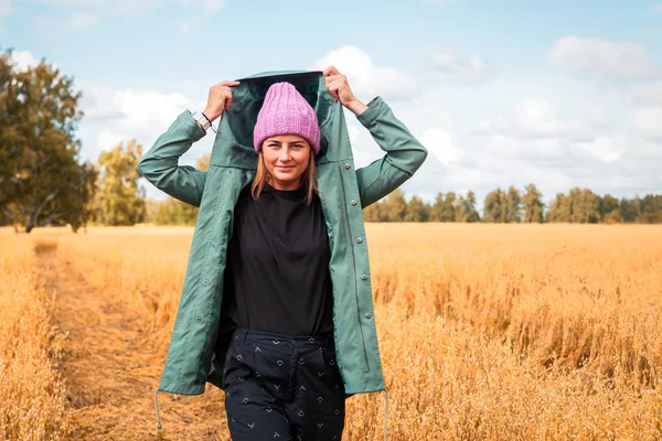 Young woman in green coat, knitting hat, jeans walking, enjoying nature and sunlight in straw field. Concept of autumn  holidays at village  and live style