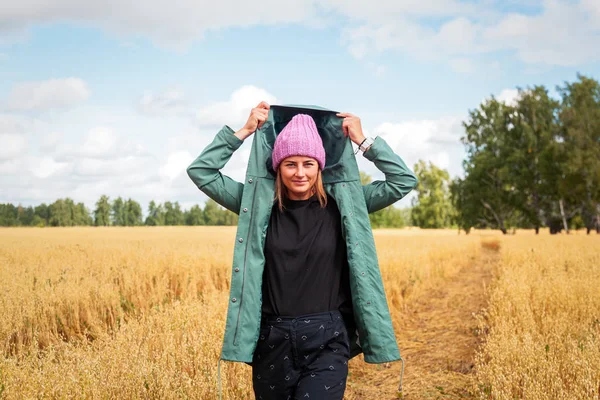 Young woman in green coat, knitting hat, jeans walking, enjoying nature and sunlight in straw field. Concept of autumn  holidays at village  and live style