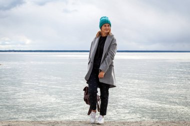 Genç güzel darkhaired kadın şapka örgü, gri bir ceket ve siyah pantolon yürüyüş ve deniz yanındaki iskelede güneşli bir sonbahar günde rahatlatıcı fotoğrafı açık atmosferik yaşam tarzı  