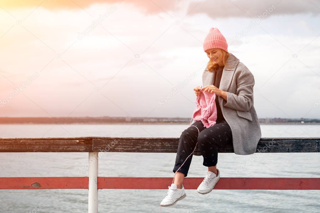 Young  married woman dressed in trendy  coat knitting a pink hat with knitting needles  against  sea  in sunny autumn day . Freelance creative working and living concept