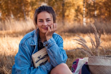 Temiz havada piknik: bir kot ceket ve elbise genç bir kadında doğa, pozlar sahiptir ve bir kitap bir arka plan manzara ve sarı sonbahar alanı ağaçlarının arkasında tutar. Livestyle ve sonbaharda açık rekreasyon kavramı