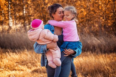 Genç bir kadın anne bir kot ceket ve elbise kızları holding ve doğanın keyfini, poz ve onlar için bir arka plan manzara ve sarı sonbahar alanı ağaçlarının arkasında konuşuyor. Livestyle ve aile açık rekreasyon kavramı 