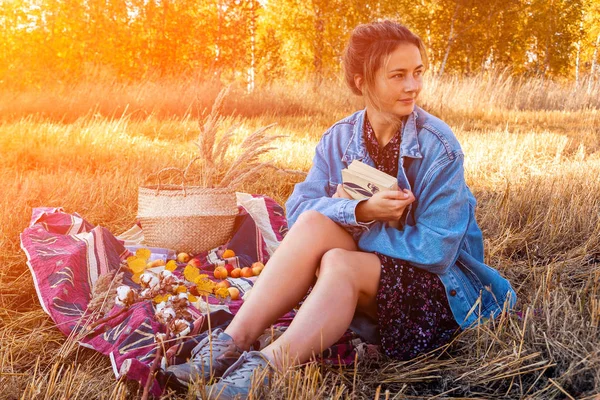 The concept of livestyle and family outdoor recreation in autumn. Picnic in the fresh air: a young woman in a denim jacket and dress hold books on plaid with a picnic basket, apples, wine. On the  background autumn  field.