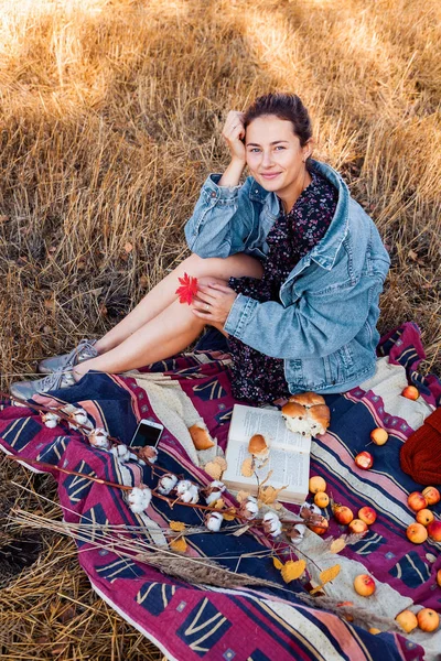 The concept of livestyle and family outdoor recreation in autumn. Picnic in the fresh air: a young woman in a denim jacket and dress enjoying nature. plaid with a picnic basket, apples, wine. On the  background autumn  field.