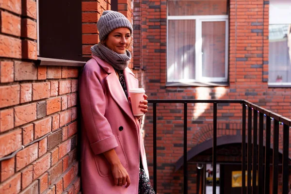 Young attractive lady in blue knitting hat and pink coat hold coffee to go . Smiling beautiful hipster happy woman in city street, wearing stylish clothes. Autumn trend, urban stydent style.