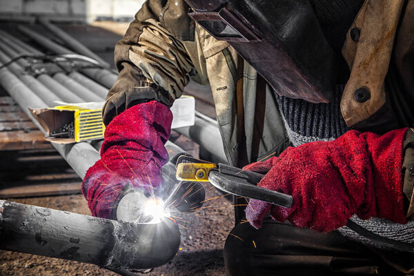 Close up welder weld metal pipe at construction site. A  builder in brown uniform, welding mask and welders leathers, weld metal with a arc welding machine, blue sparks fly to the sides