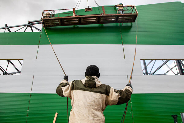 The builders are building a building of metal structures at a height in the construction cradle, in the foreground is a builder holding ropes against a blue sky. Violation of safety at a construction site