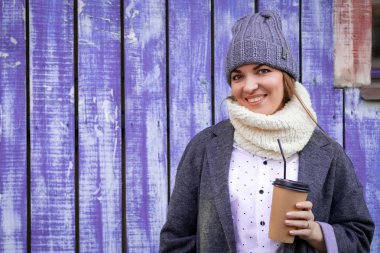 Bir gri örgü şapka, gri ceket ve bluesses genç bir kadın şehir, pozlar ve içecek kahve mavi duvar sahiptir. Livestyle ve sonbaharda açık rekreasyon kavramı