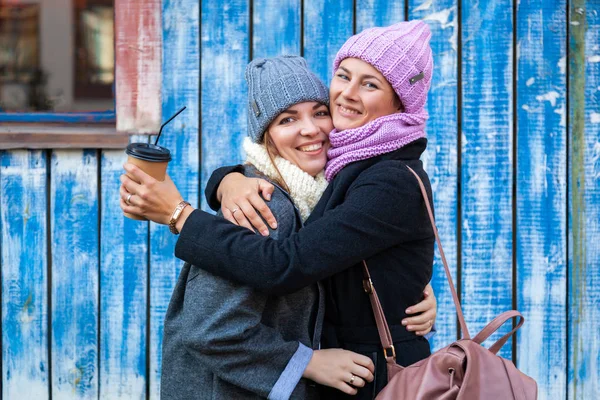 A young women friends in a stylish clothes   hug, chat  and drink coffee to go around blue wall. The concept of livestyle and outdoor recreation in autumn
