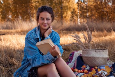 Temiz havada piknik: bir kot ceket ve elbise genç bir kadında doğa, pozlar sahiptir ve bir kitap bir arka plan manzara ve sarı sonbahar alanı ağaçlarının arkasında tutar. Livestyle ve sonbaharda açık rekreasyon kavramı