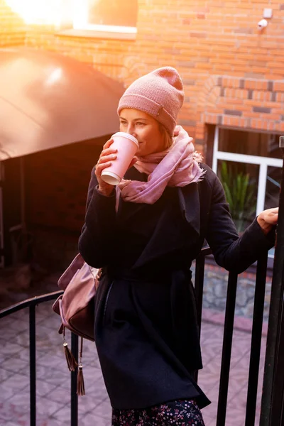Young attractive lady in pink knitting hat and black coat drink coffee to go . Smiling beautiful hipster happy woman in city street, wearing stylish clothes. Autumn trend, urban stydent style.