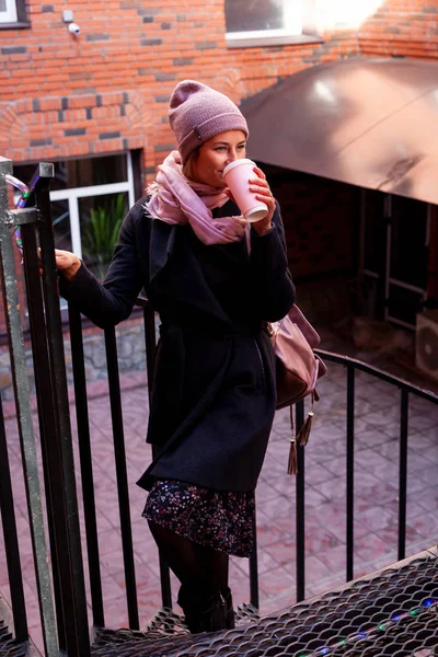 Young attractive lady in pink knitting hat and black coat drink coffee to go . Smiling beautiful hipster happy woman in city street, wearing stylish clothes. Autumn trend, urban stydent style.